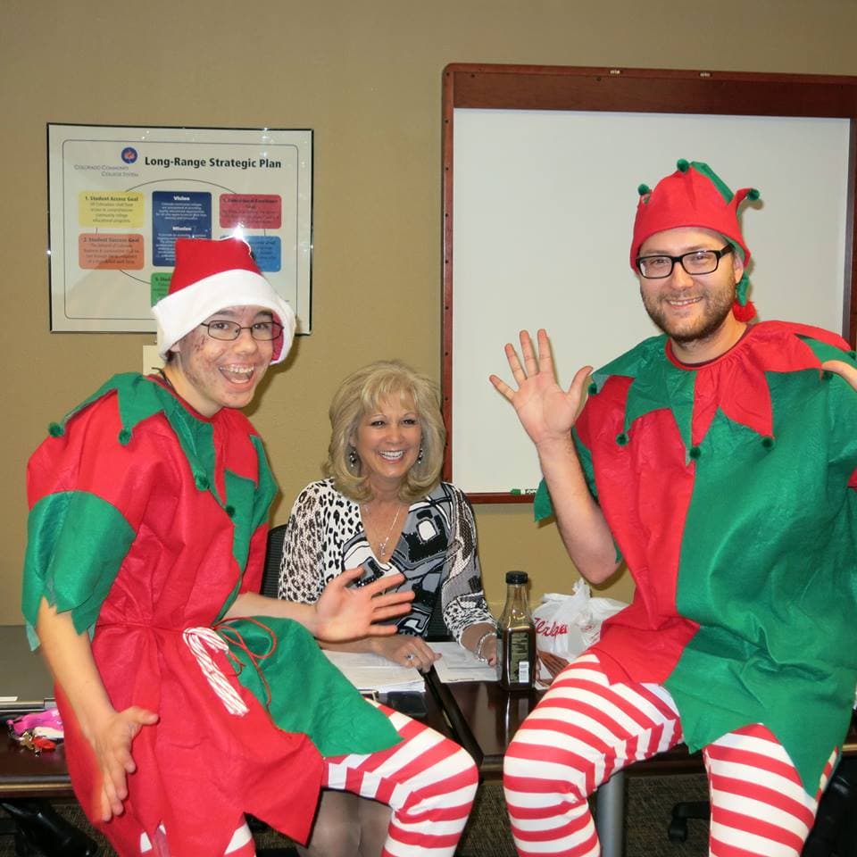 Two students in elf costumes wave beside President Patty Erjavec at a meeting table