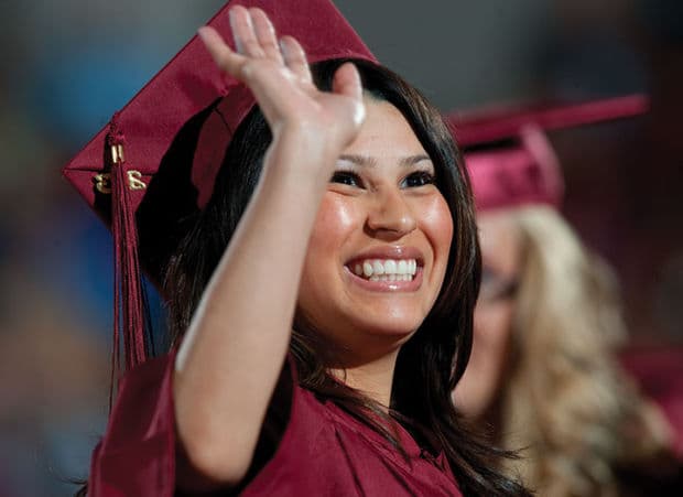 A smiling PCC graduate in a maroon cap and gown waves during commencement