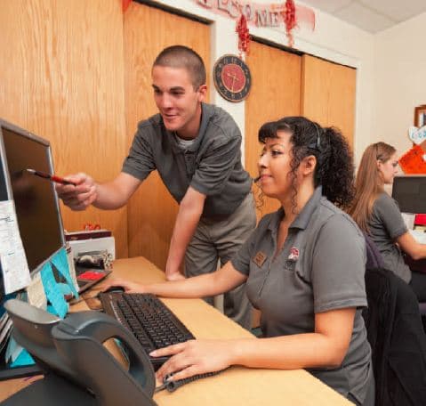 A young man points to a computer screen as a student in a PCC polo shirt works at the keyboard